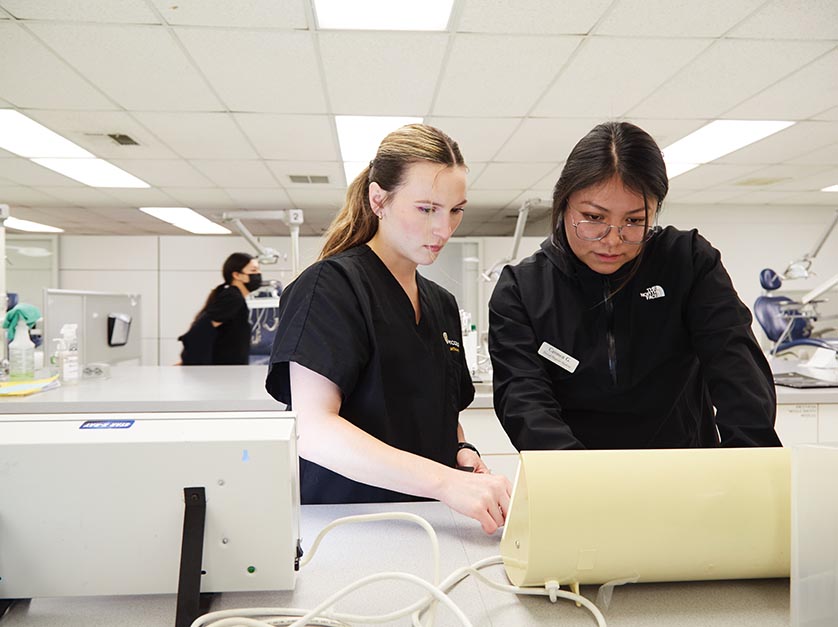 Two female Dental Hygiene students stand side-by-side in the dental clinic and evaluate a set of x-rays.
