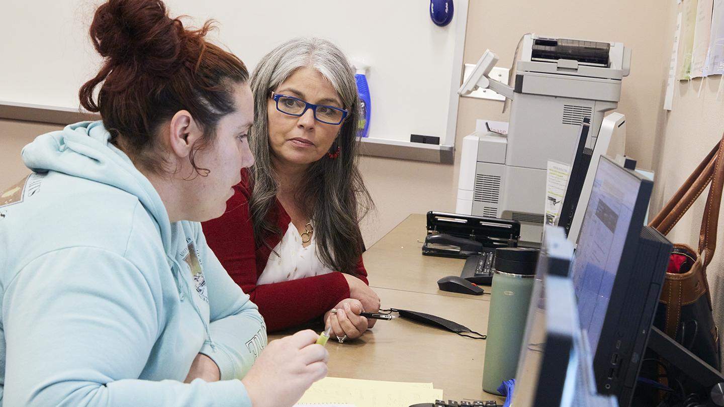 Female professor, seated at a computer table next to a female student, looks at the student to see if she understands what is on the papers in front of them.