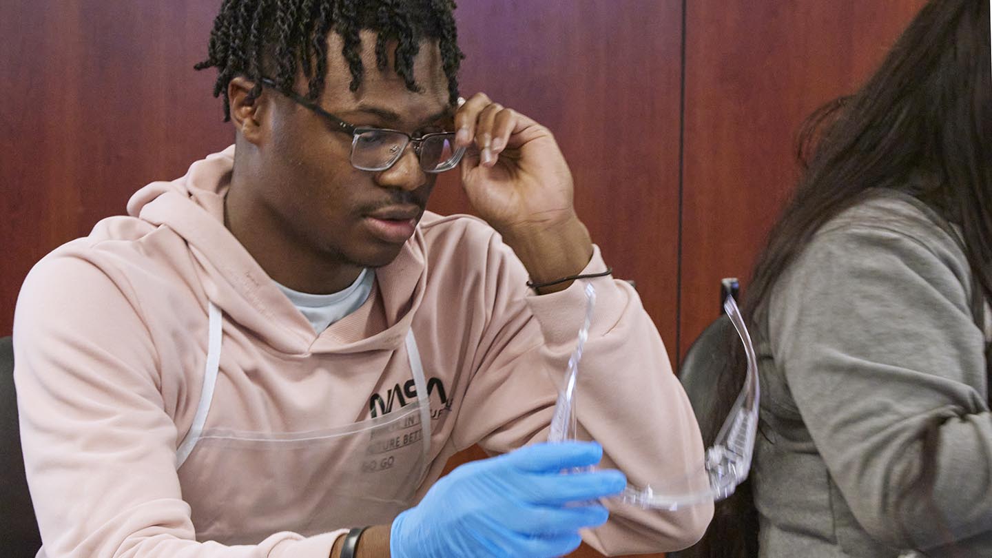 Male student, sitting at a lab table, adjusts his eyeglasses and holds safety goggles with his other hand.